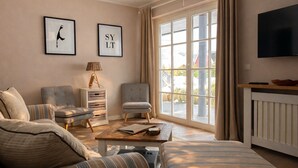 A living room with a sofa, two armchairs, a coffee table, and a TV. There are two framed pictures on the wall and a window with curtains. - Beautiful apartment with large south-facing terrace (Sankt Peter-Ording)