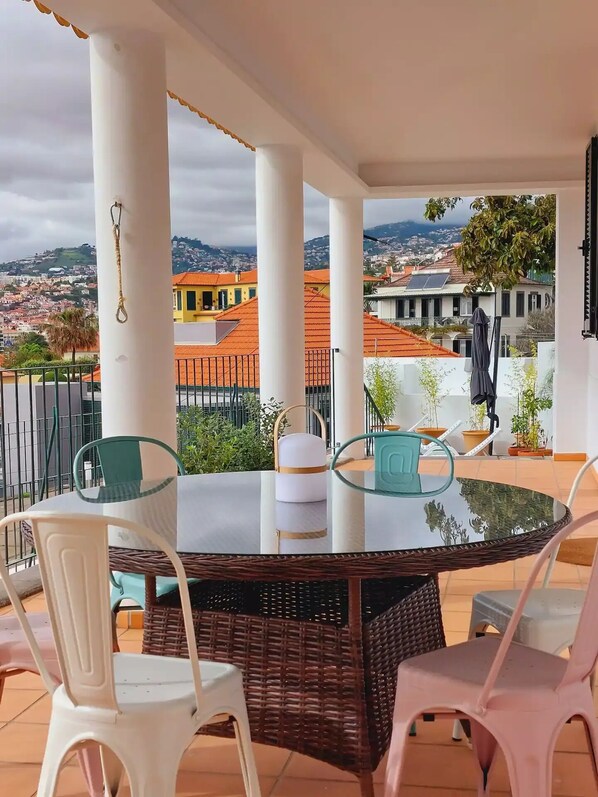 A round glass table with wicker chairs on a balcony overlooking a cityscape with red-roofed buildings.