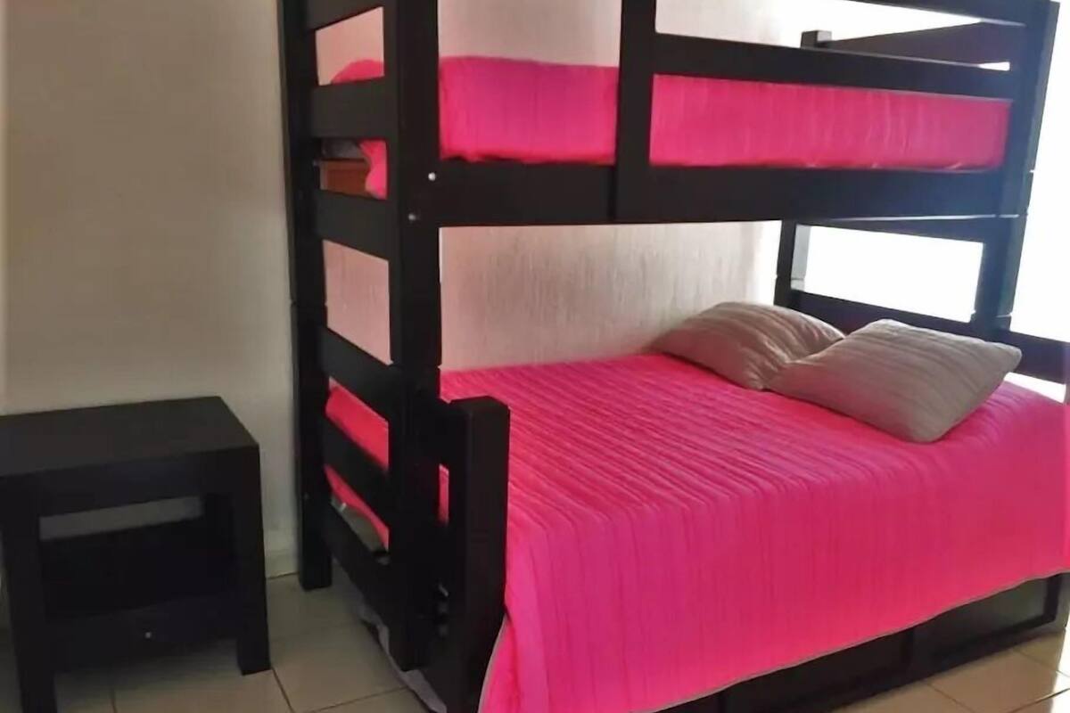 Uma cama beliche com colcha e almofadas rosa, um criado-mudo preto pequeno e uma janela com cortinas brancas.