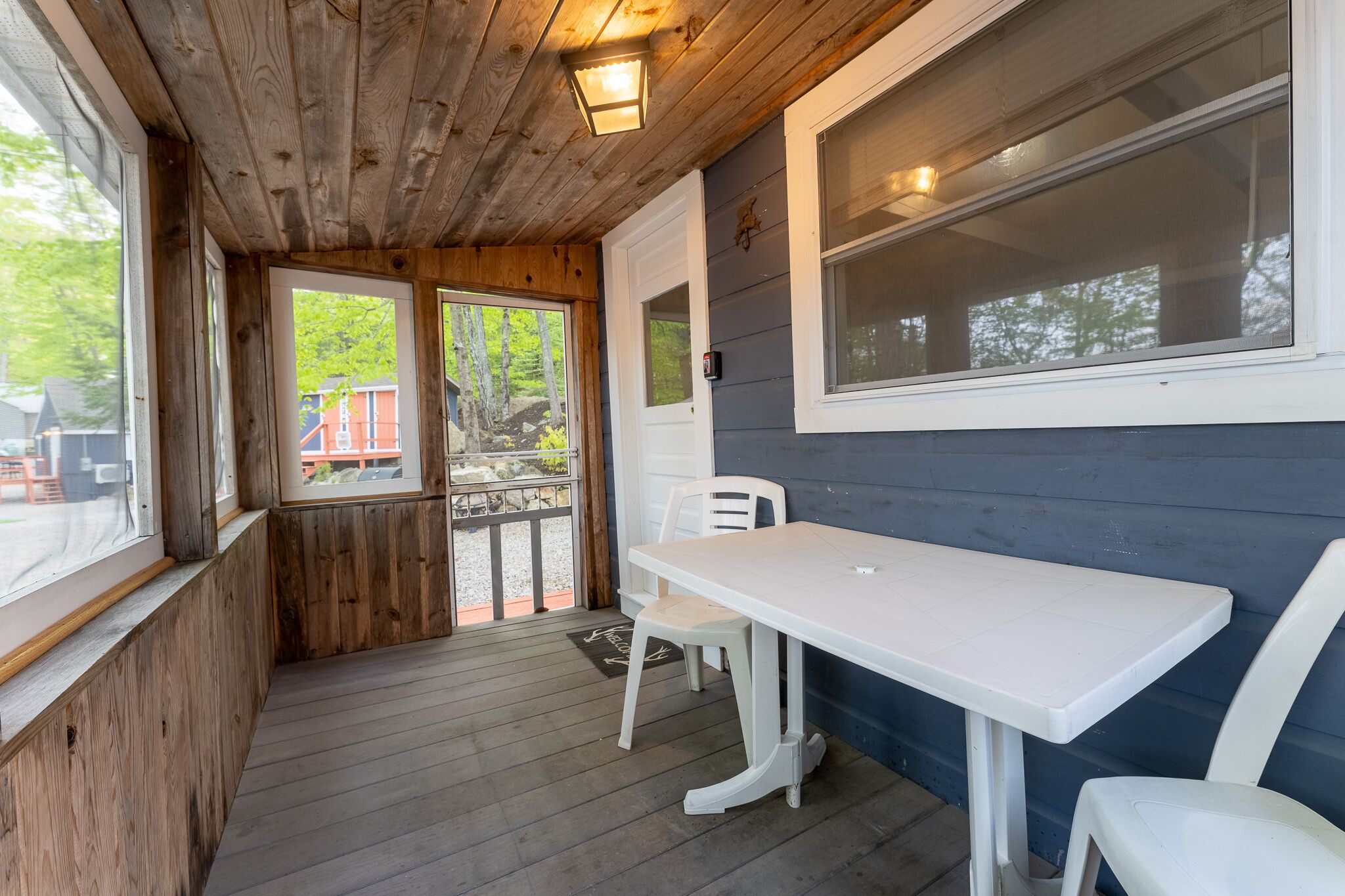 Une terrasse en bois avec une table et des chaises blanches, une grande fenêtre et une porte donnant sur l’extérieur.