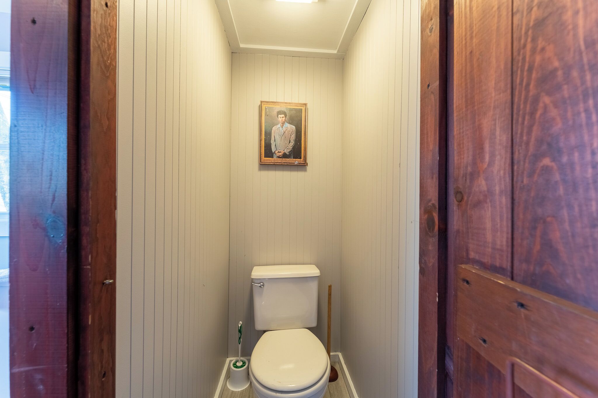 Une petite salle de bain aux murs en bois, avec des toilettes blanches, une brosse de toilette verte et un tableau encadré accroché au mur.
