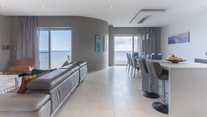 Una sala de estar moderna con un sofá gris, un comedor con taburetes de bar y un ventanal con vistas al mar.