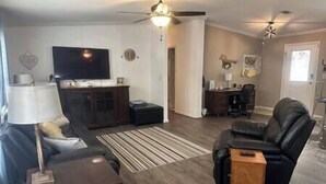 A living room with a sectional sofa, a small wooden side table, a television on a cabinet, a ceiling fan, and a rug. - The K Way, The K Way (Ocala)