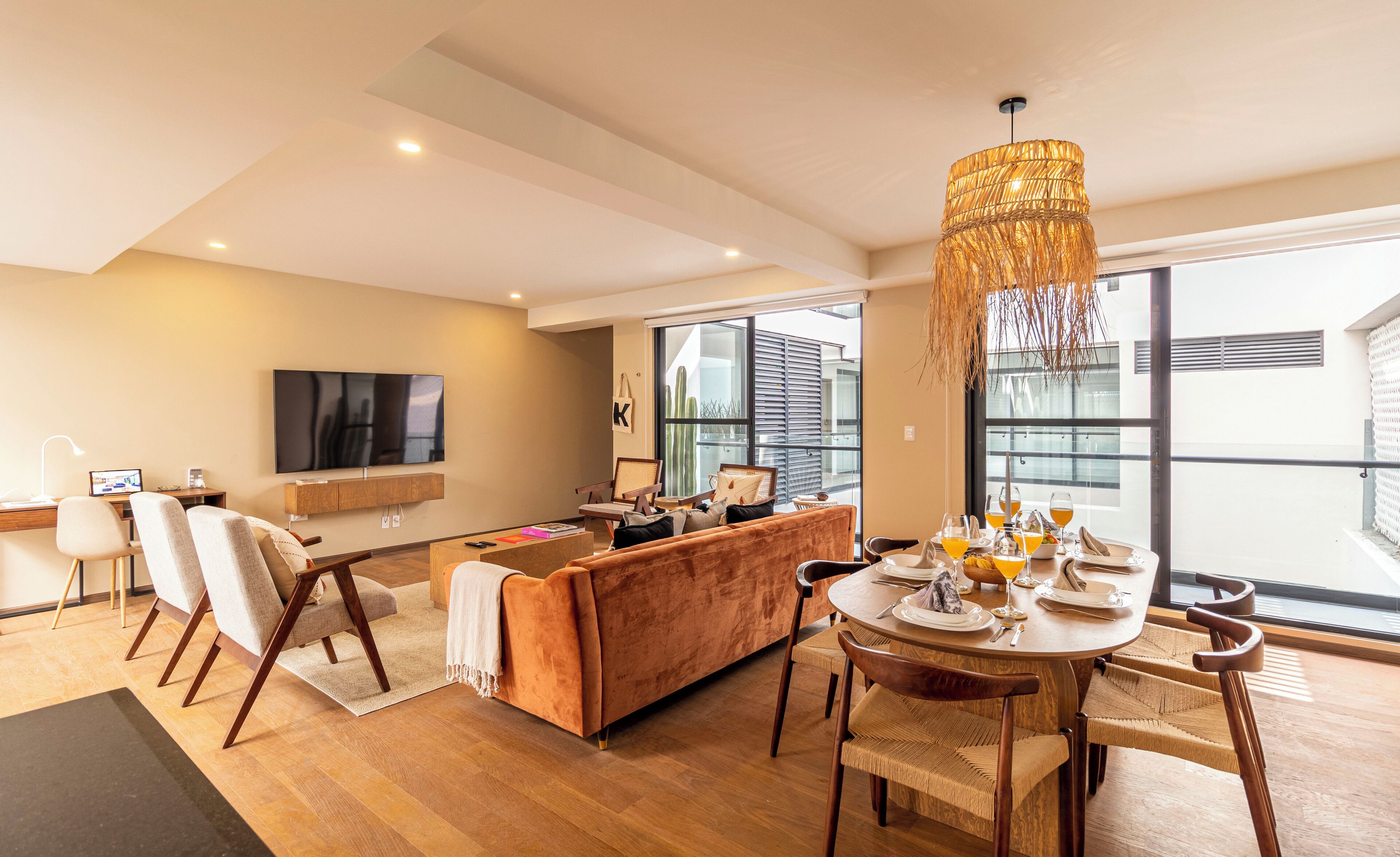 A modern living room with a sofa, dining table, and chairs, a TV mounted on the wall, and a large window providing natural light.