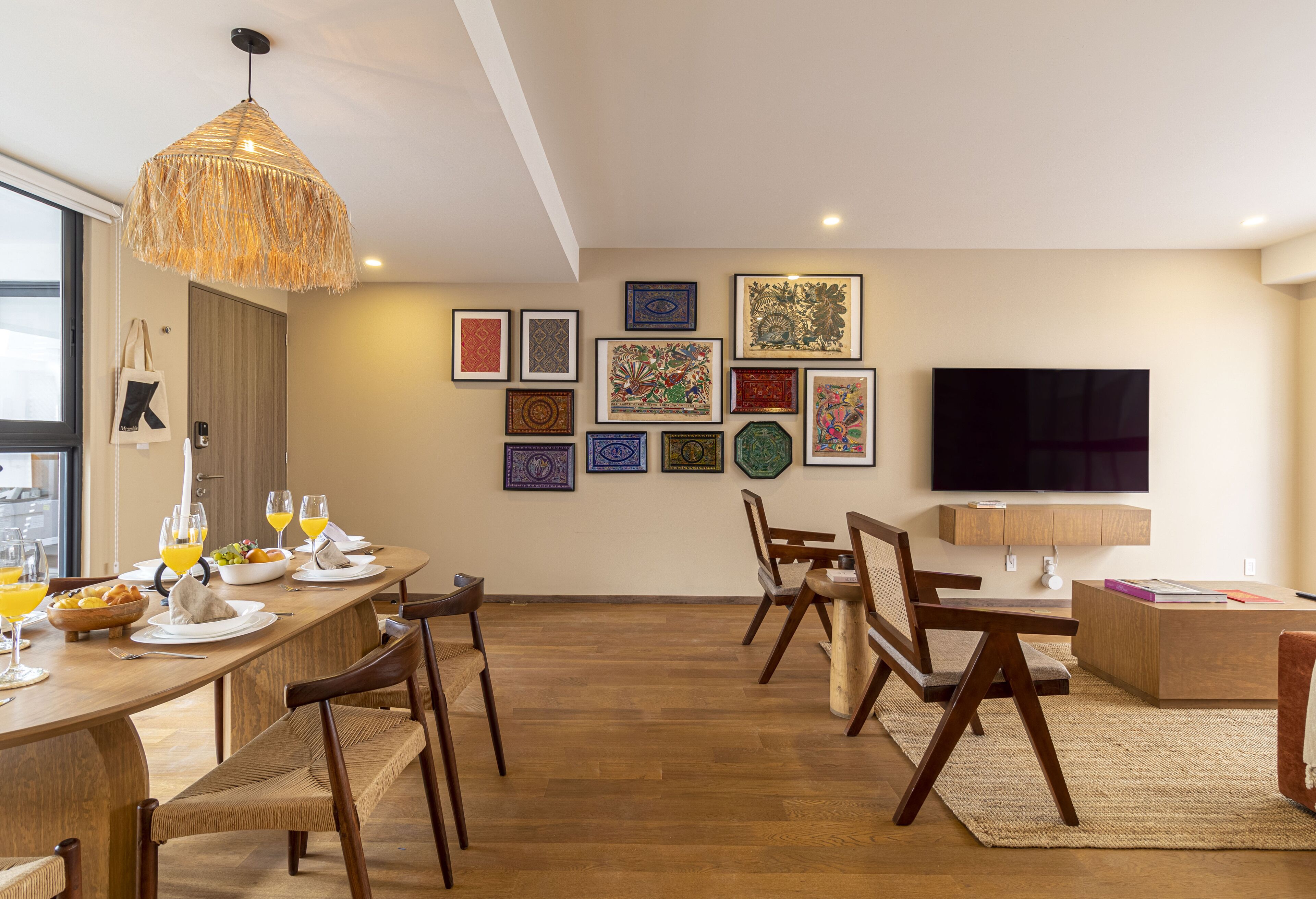 A dining area with a long wooden table set for a meal, surrounded by chairs. There is a television mounted on the wall and a variety of framed artworks displayed above the table.