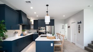 Cocina moderna con armarios azul oscuro, encimera blanca y una isla central. Hay un comedor con una silla de madera y una cesta de objetos.