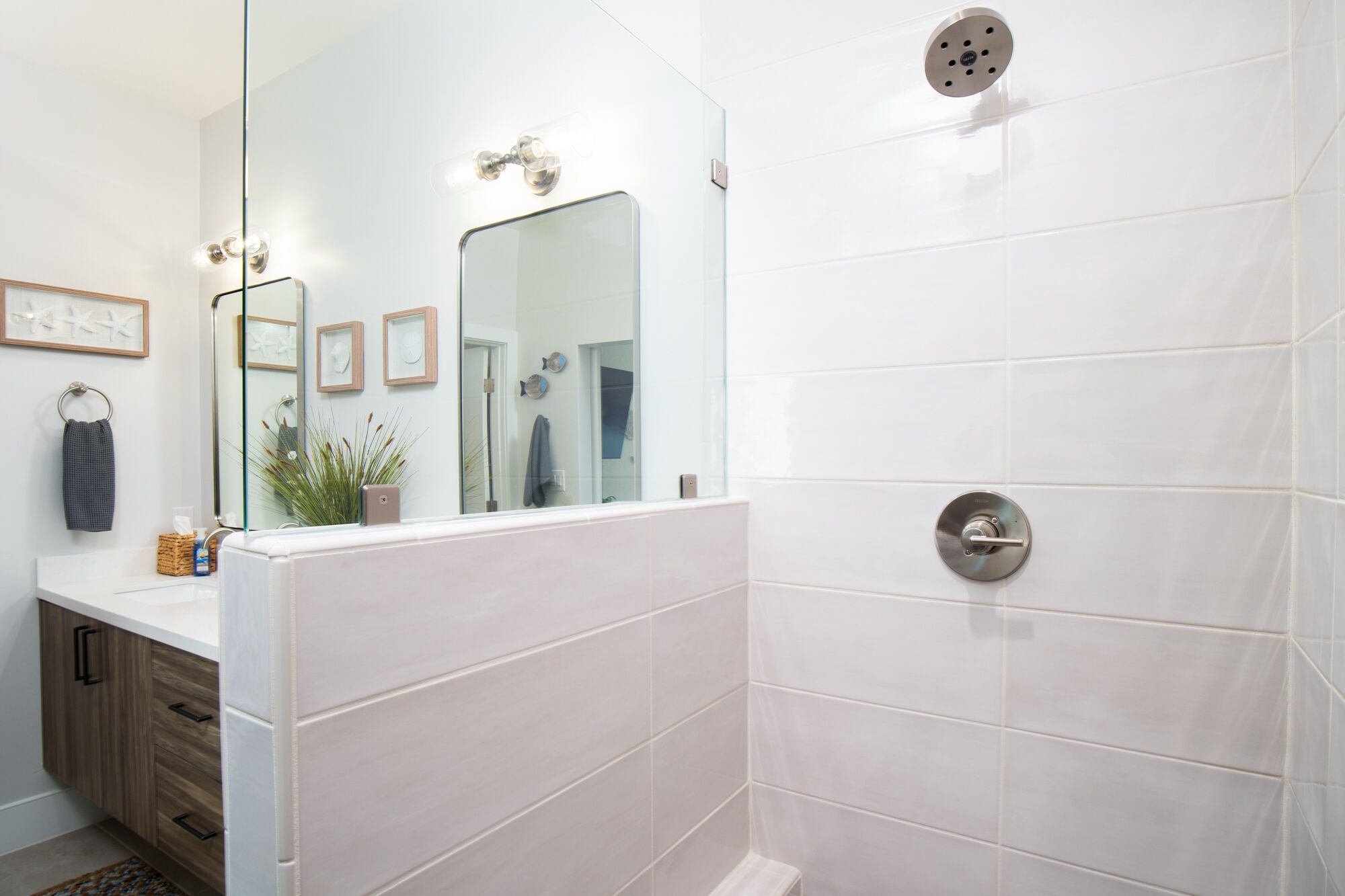 A modern bathroom with white tiles, a large mirror, a shower with a control panel, and a wooden cabinet with drawers.