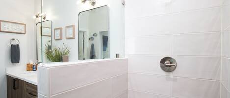 A modern bathroom with white tiles, a large mirror, a shower with a control panel, and a wooden cabinet with drawers.