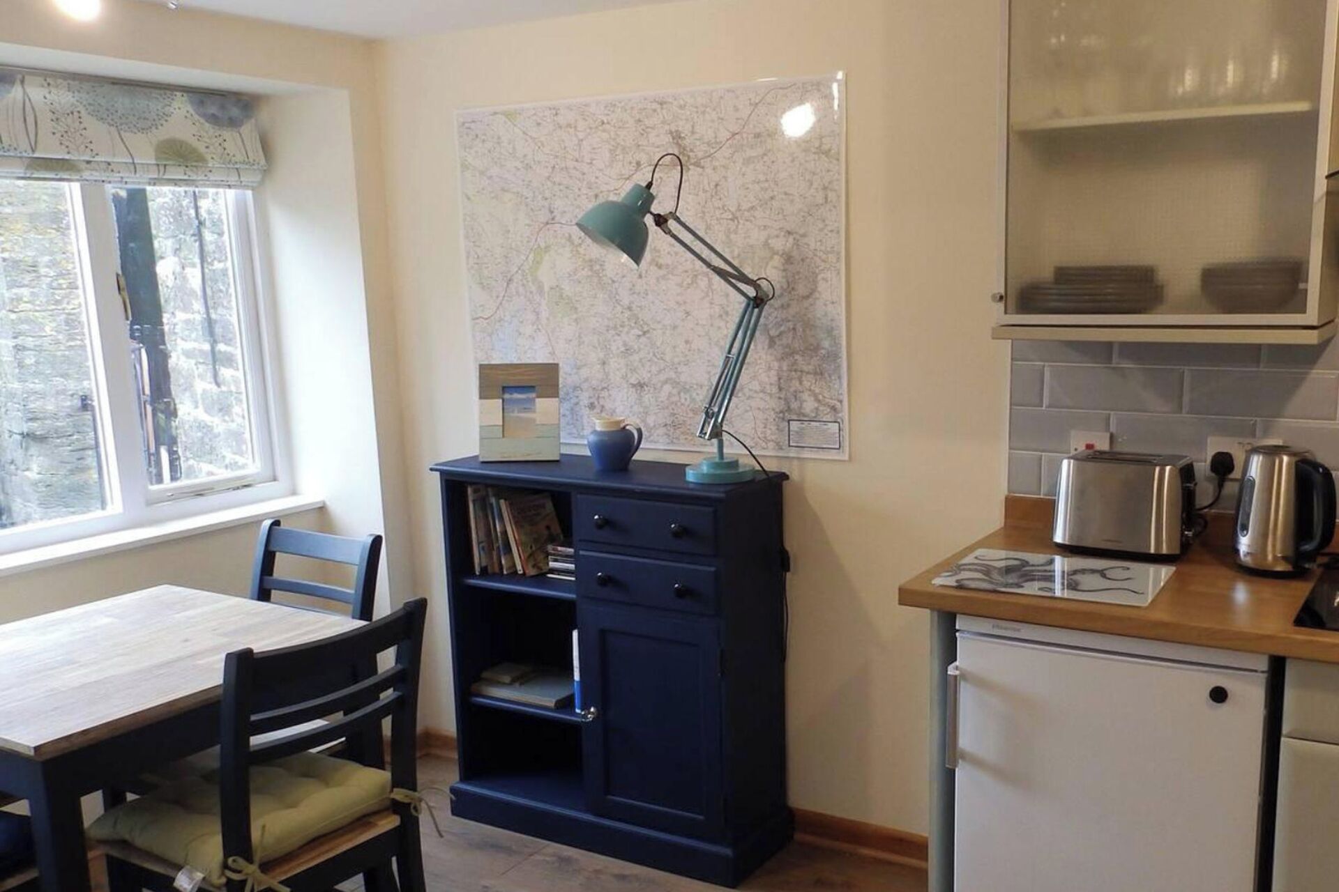 A compact kitchen with a small refrigerator, a toaster, and a kettle on the counter. There's a desk with a chair and a map on the wall.