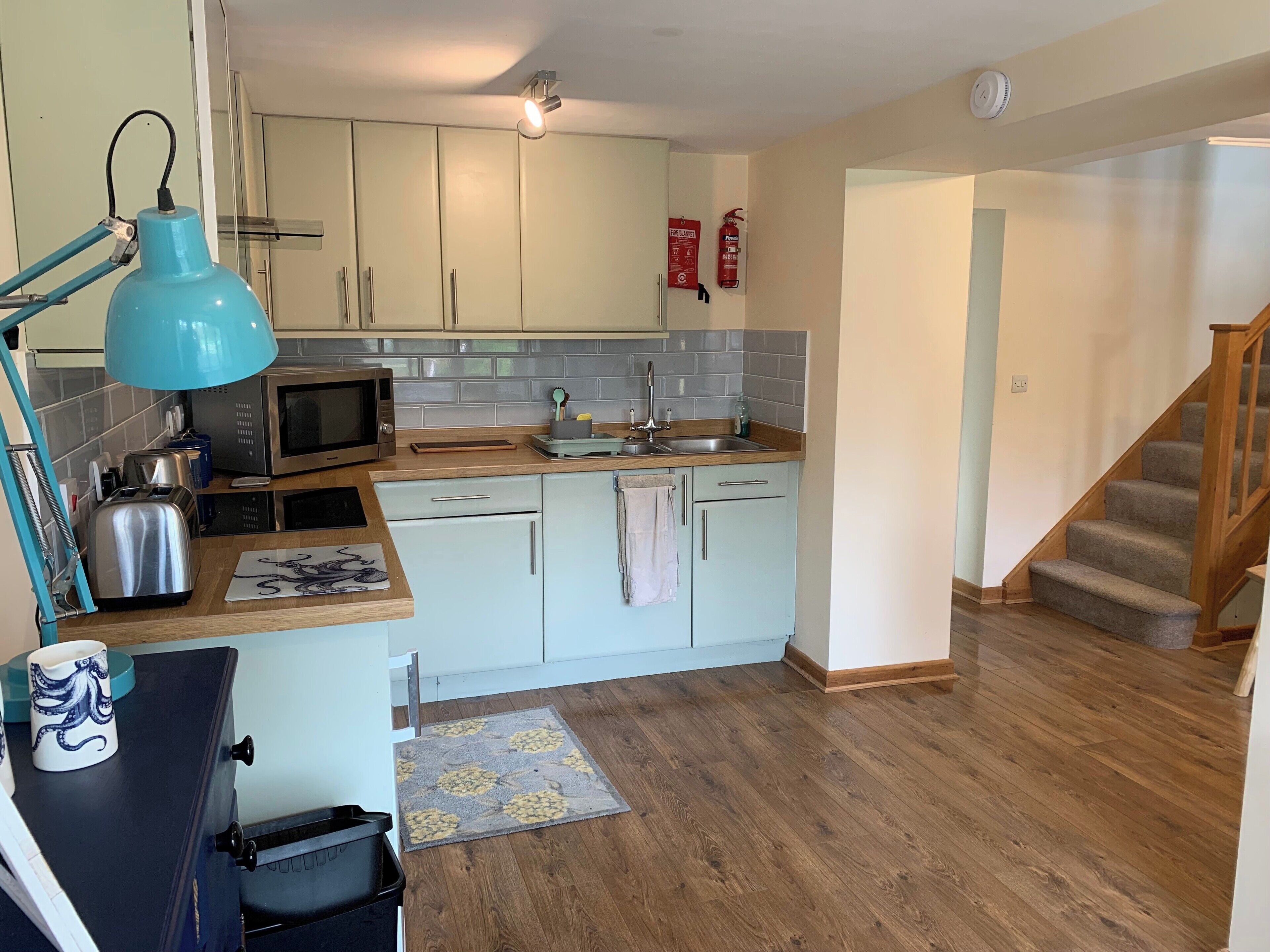 A kitchen with white cabinets, a microwave, a toaster, and a sink. There is a blue pendant light, a fire extinguisher, and a wooden staircase.