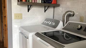 A laundry area with a front-loading washer and dryer, a shelf with various cleaning products, and a ventilation system.