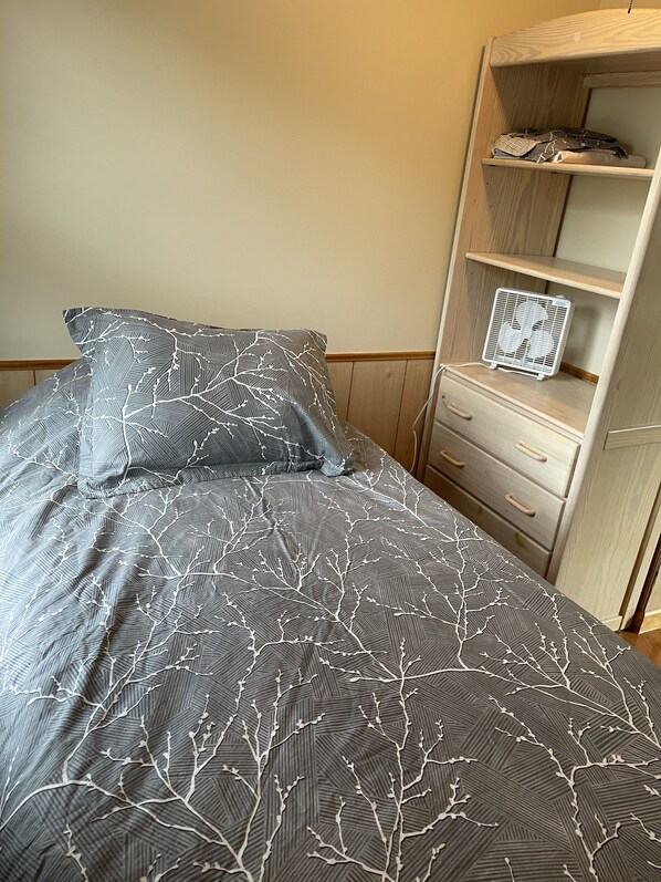 A bed with a grey patterned bedspread and pillows, a white bedside table with drawers, and a wooden wardrobe with shelves.