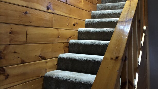 Una escalera de madera con barandilla, peldaños alfombrados y un ventanal con persianas.
