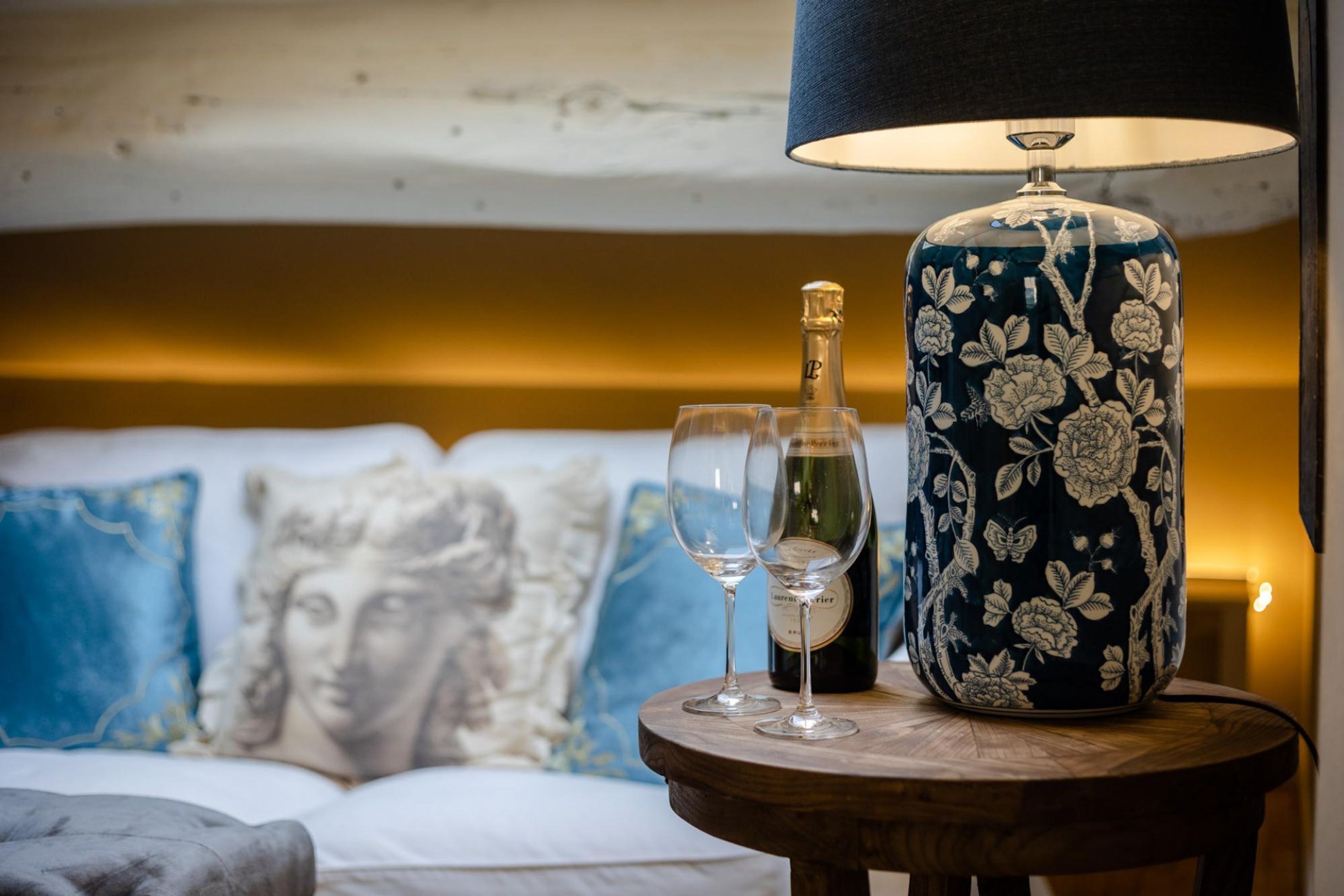 A room with a bed, a round wooden table with a bottle and two glasses, a lamp, and a sofa with patterned cushions.