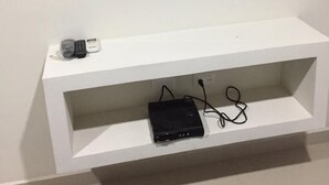 Uma televisão de tela plana fixada em uma parede branca, acima de um móvel branco que contém um laptop e um controle remoto.