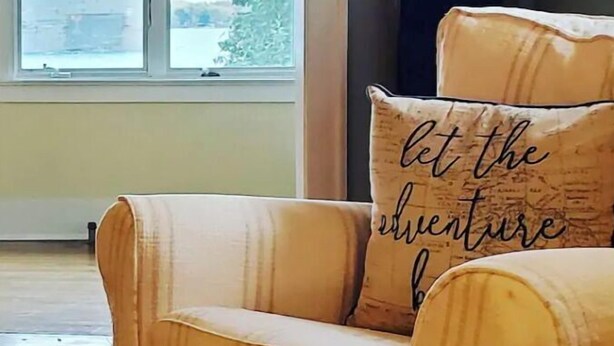 A room with a large window, a sofa with a cushion that says "let the adventure begin," and a television on the wall.