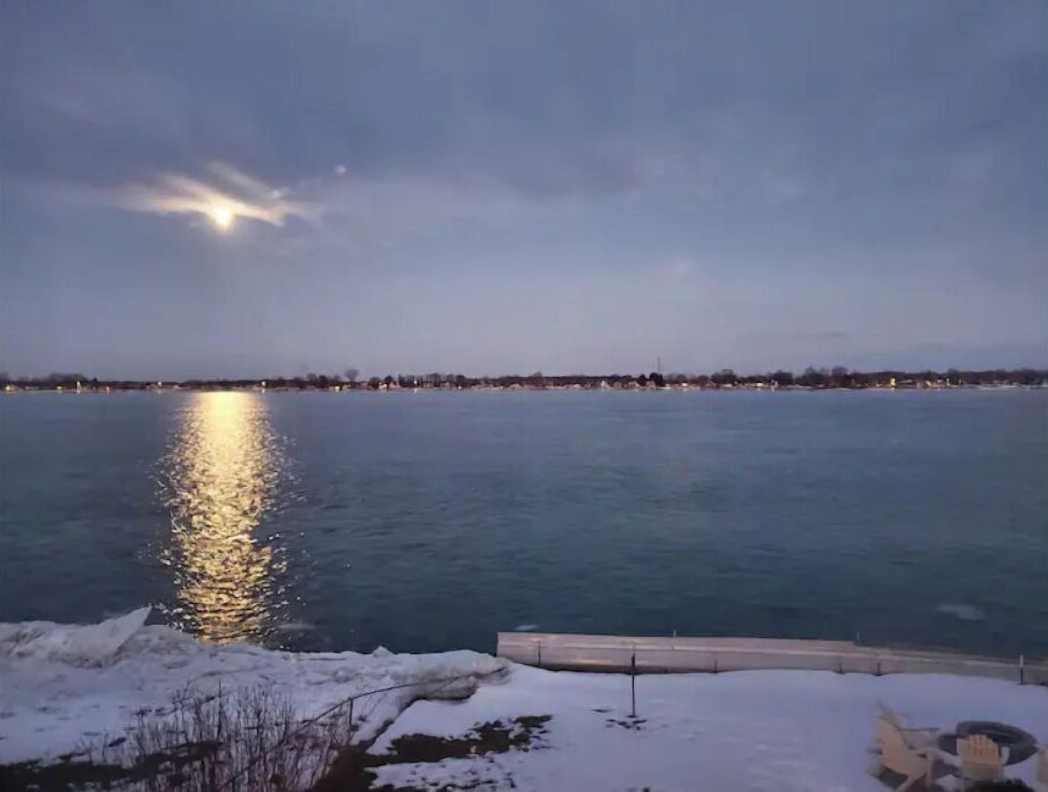 A waterfront view with a calm body of water reflecting lights, a snow-covered foreground, and a distant shoreline with buildings.
