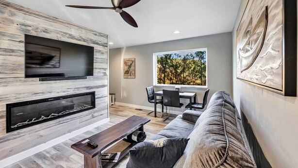A modern living room with a fireplace, a ceiling fan, and a large window with a view of trees. - CONDO 5-Sister Bay Gem-Steps to the Beach (Sister Bay)