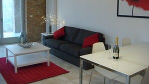 Una sala de estar moderna con un sofá gris, cojines rojos, una mesa de centro blanca y una mesa de comedor con una botella de champán.