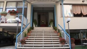 L’entrée d’un hôtel, sur laquelle on peut lire l’inscription « HJASMINE ». Des marches mènent à l’entrée, de part et d’autre, on trouve des plantes en pot et des garde-corps bleus.