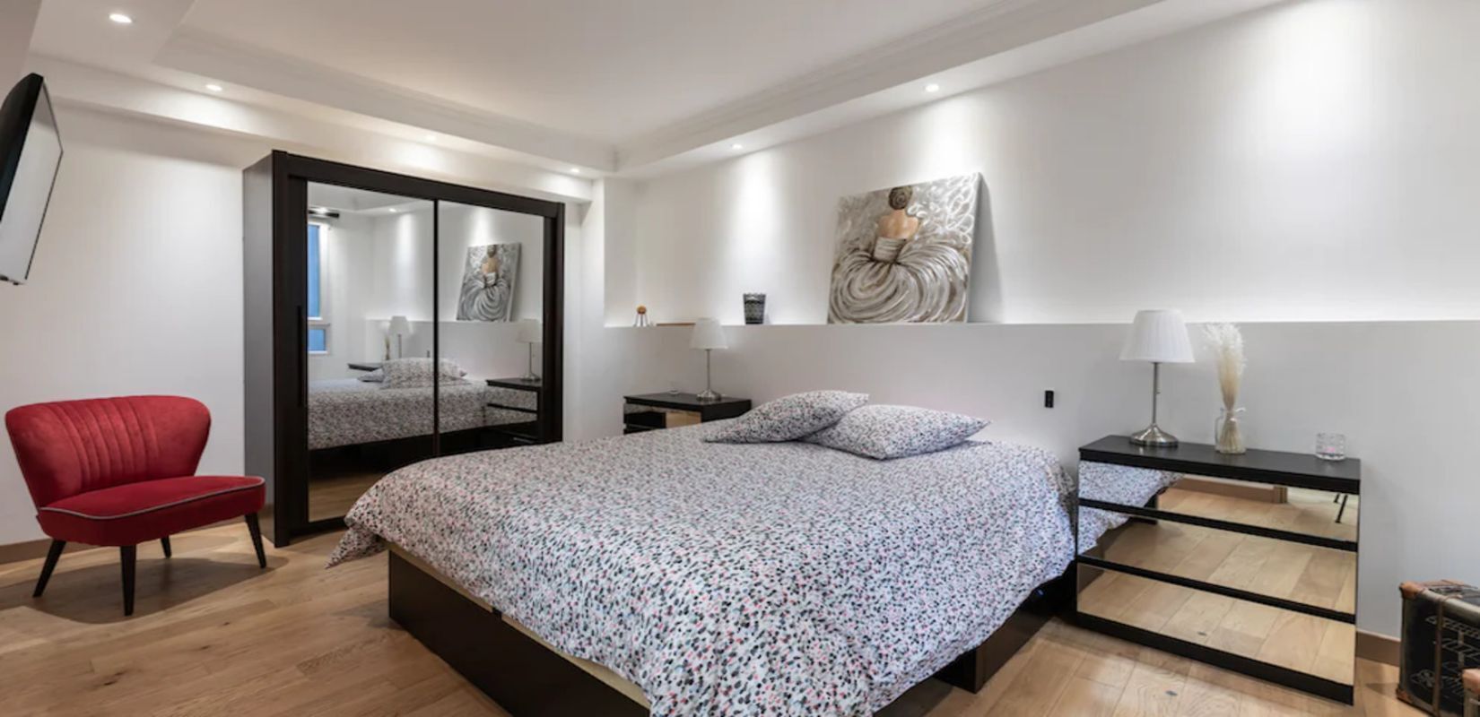 A modern bedroom with a large bed, a red chair, a mirror, and a bedside table with a lamp.