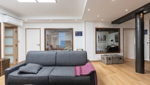 Un salon moderne avec un canapé gris, un plancher en bois et un grand miroir qui reflète la pièce. On y voit des valises et une lampe posées sur le sol.