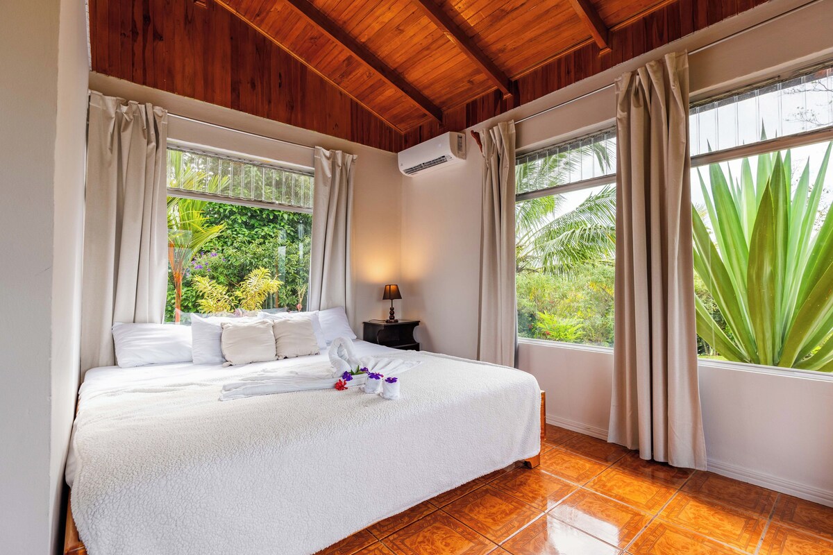 A neatly made bed with white linens and a towel, a bedside lamp, and a view of greenery through large windows.