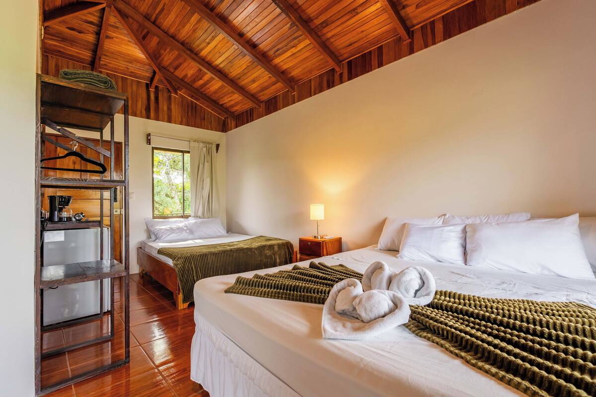 A hotel room with two beds, a wooden ceiling, a small table with a lamp, and a shelf with hangers.