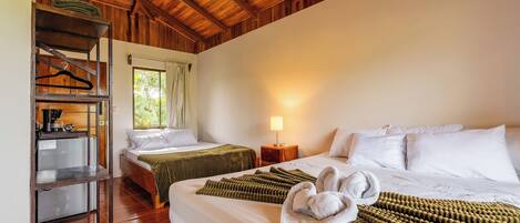A hotel room with two beds, a wooden ceiling, a small table with a lamp, and a shelf with hangers.