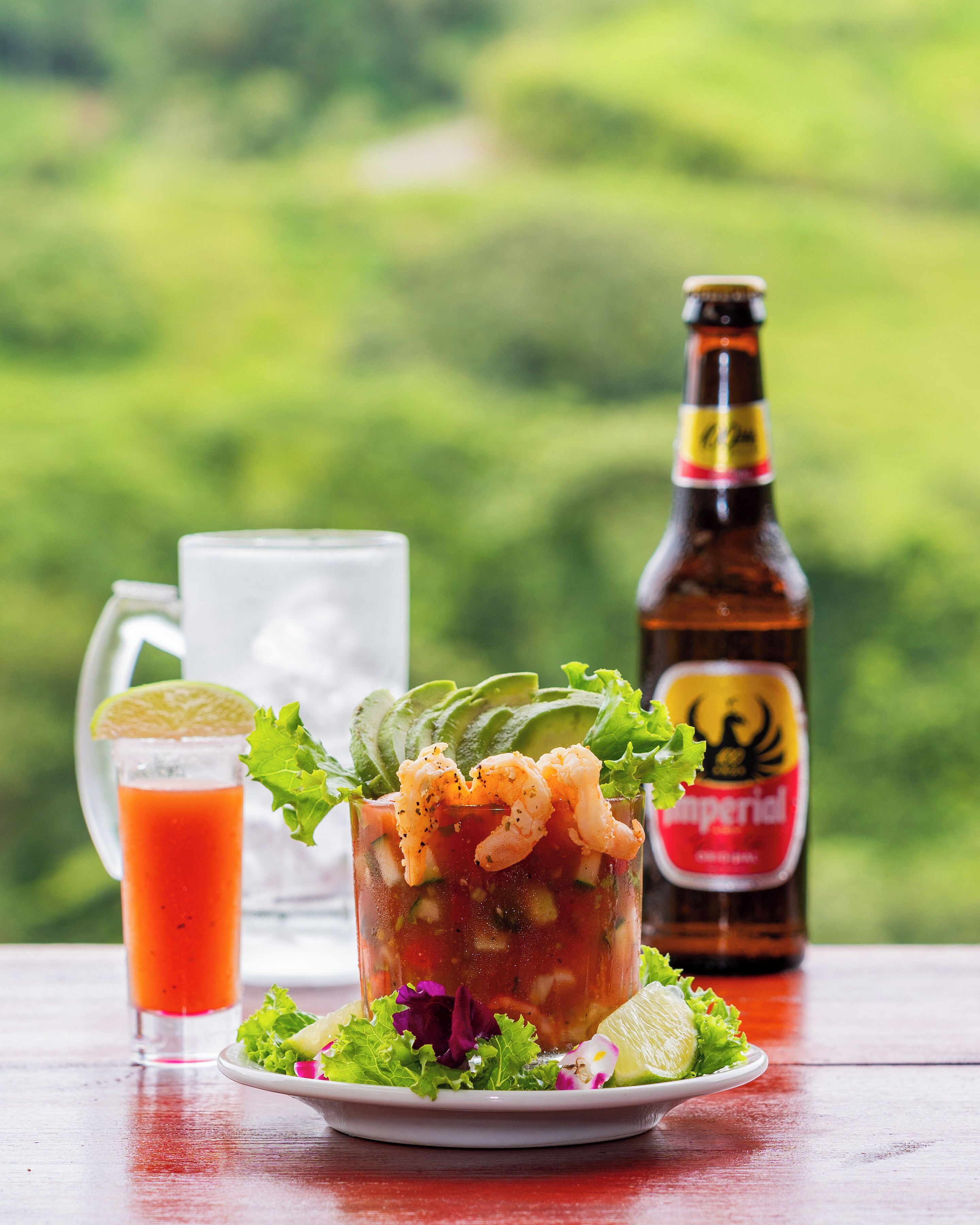 A cocktail with shrimp garnish, a bottle of beer, and a glass of juice on a table with a green landscape in the background.
