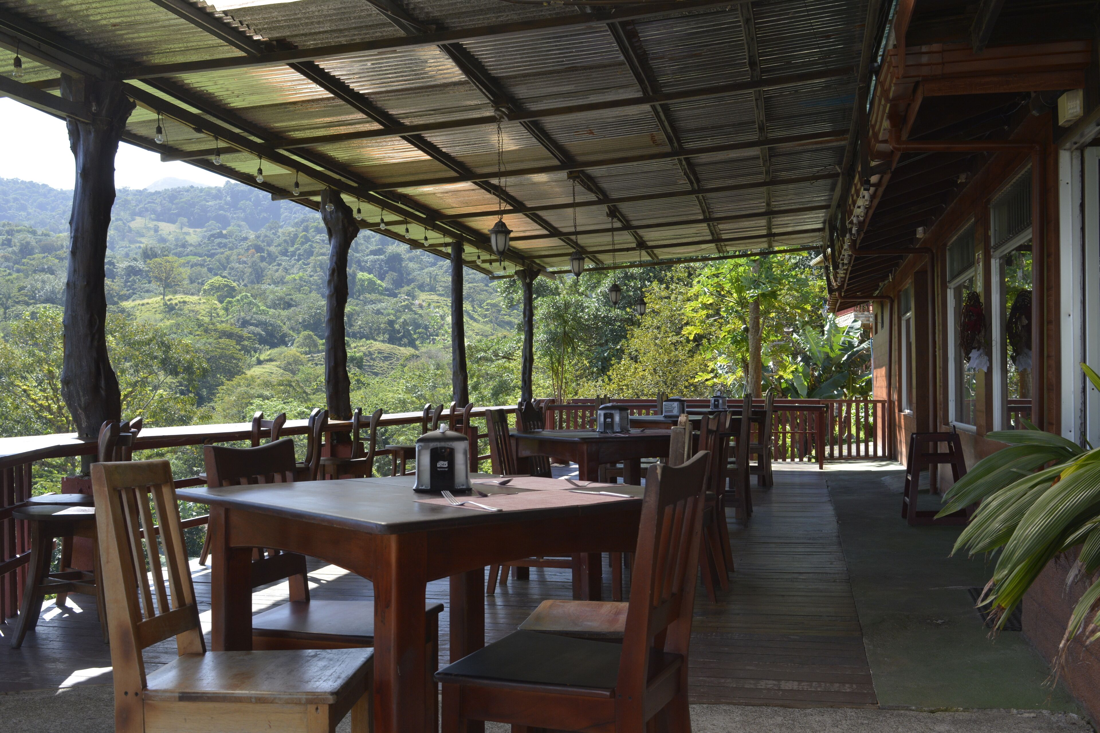 An outdoor dining area with wooden tables and chairs, surrounded by a metal roof, overlooking a lush green landscape.
