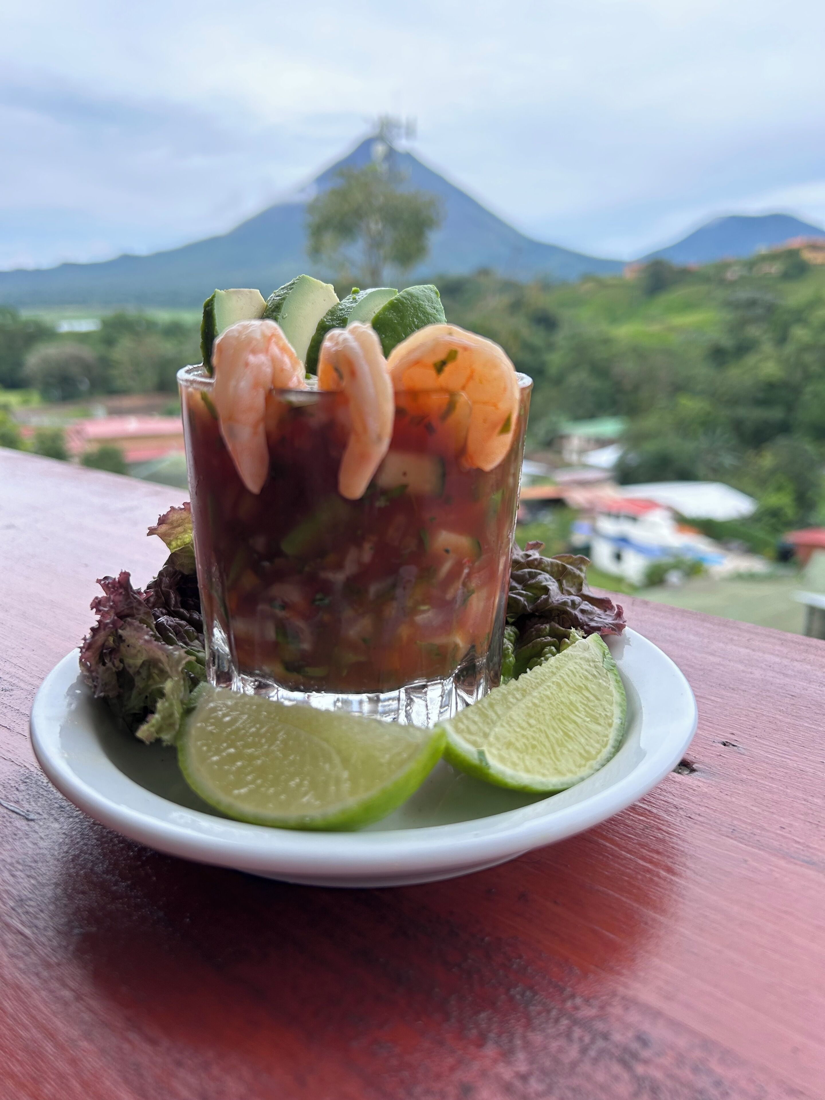 A shrimp cocktail with lime and cucumber garnish, served on a white plate with a wooden table and a scenic mountain view in the background.