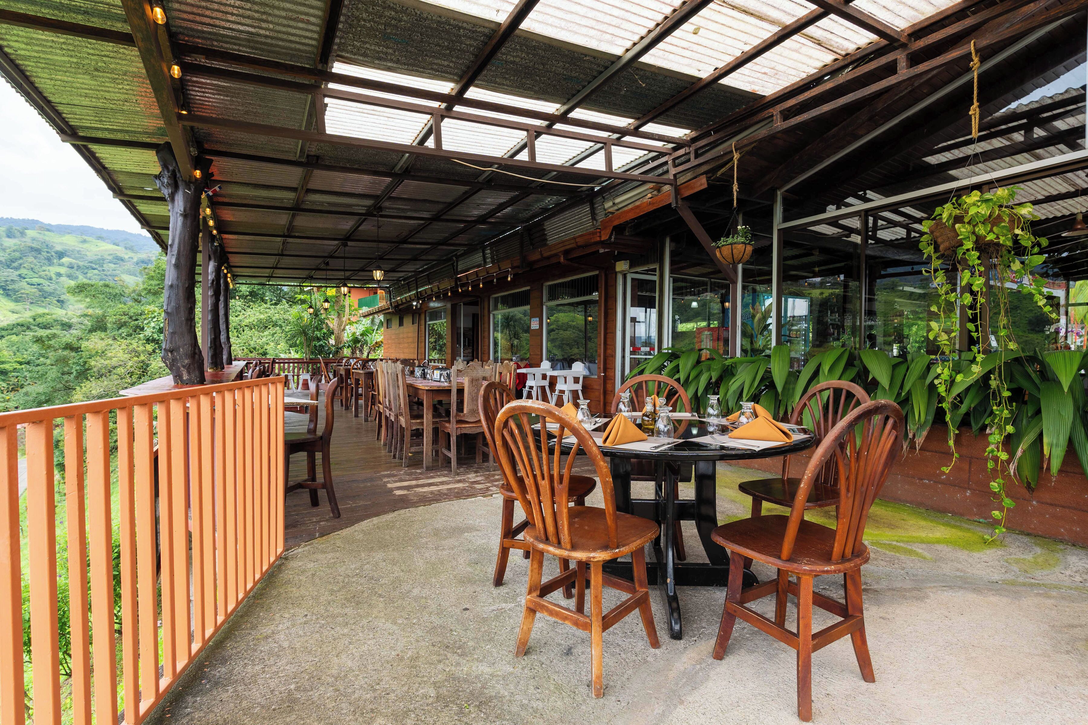 Un espace de restauration en plein air, avec des chaises et des tables en bois, entouré d’une rambarde en bois et abrité par un toit en métal sous lequel pendent des plantes.