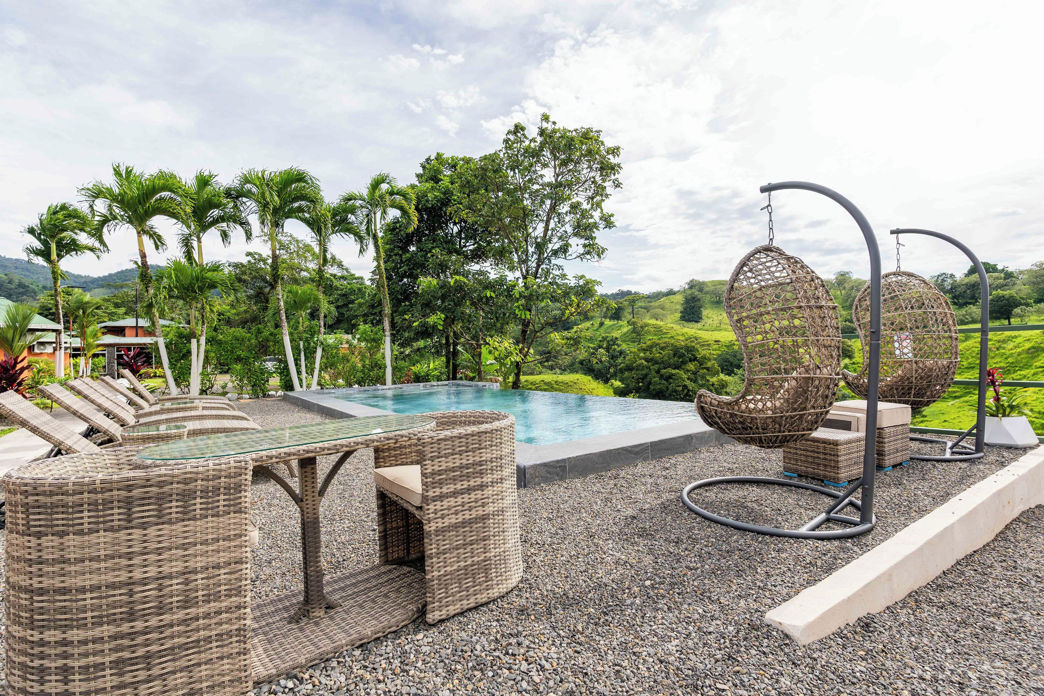 Un área junto a la piscina con mobiliario de mimbre, sillones colgantes y vistas a vegetación exuberante y colinas.