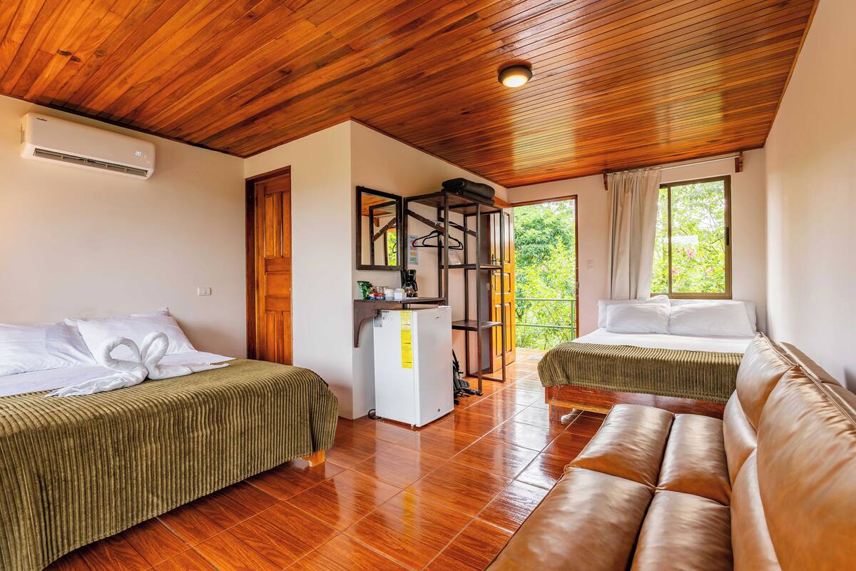 A room with two beds, a wooden ceiling, a refrigerator, and a view of greenery outside.