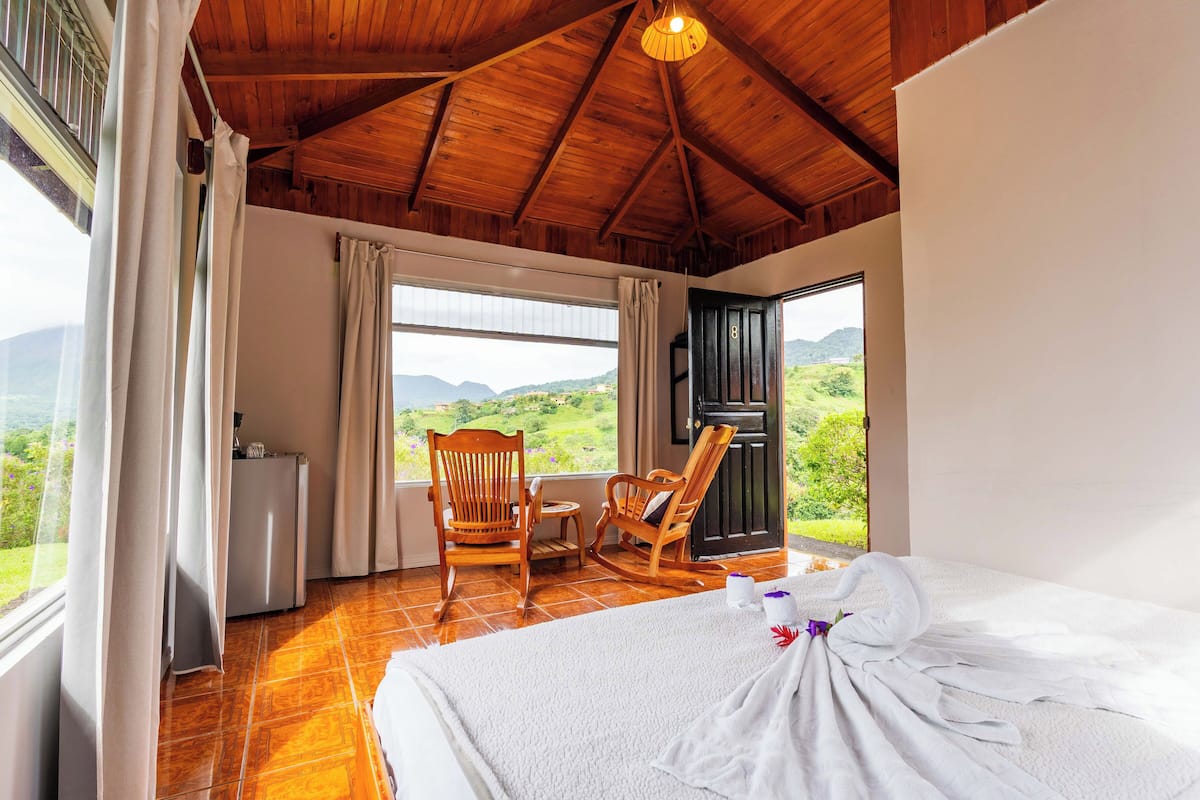 A bedroom with a large bed, a wooden rocking chair, a small table, and a view of the outdoors.