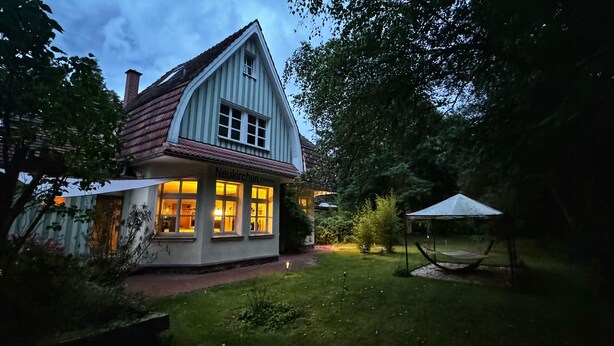 A traditional house with a sloped roof and a lit-up front window, surrounded by a well-maintained garden and a small gazebo. - Schmuckstück traumhafter ehem. Bahnhof in Alleinlage (Braunfels)