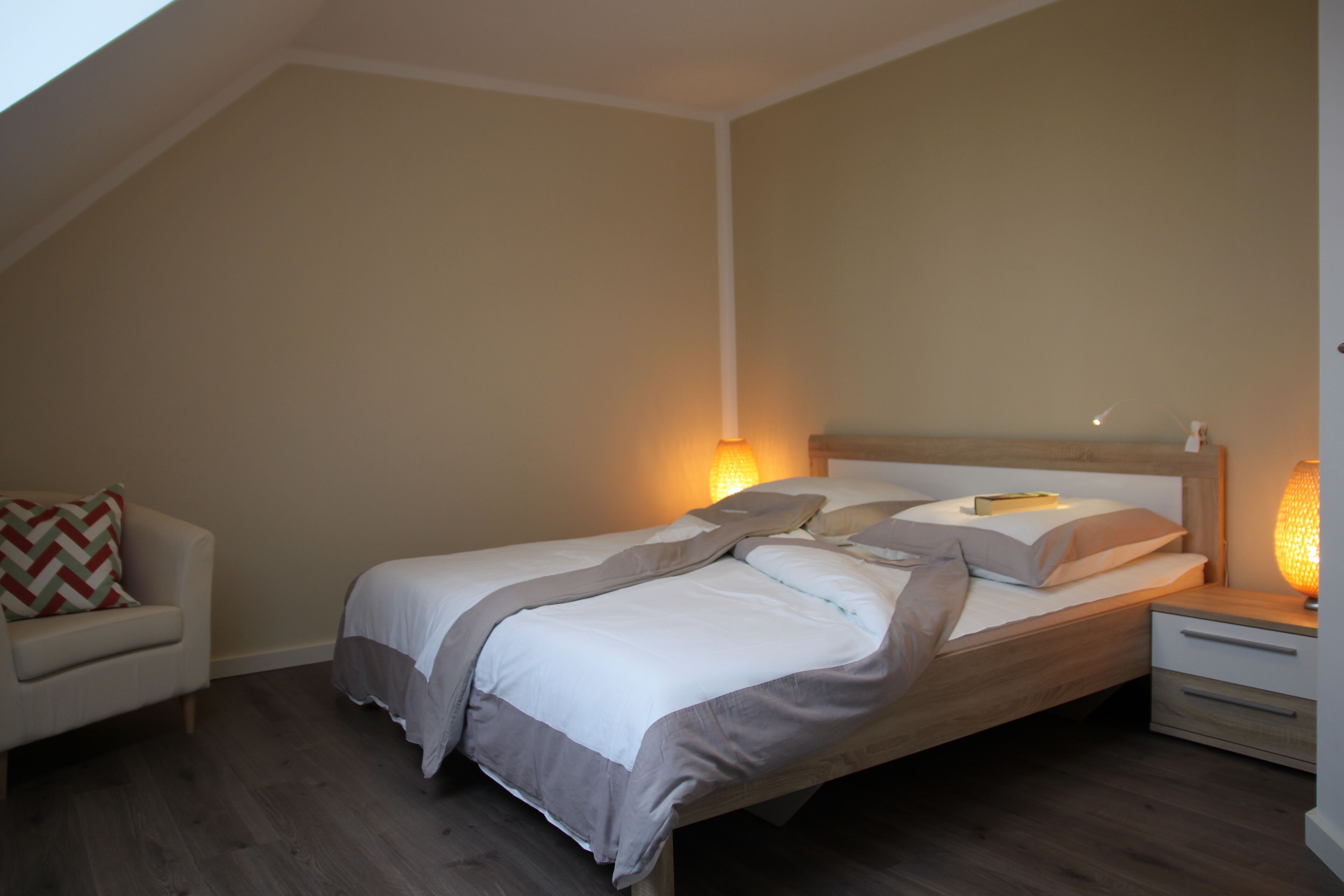 Een slaapkamer met een bed, nachtkastje, lamp en stoel.