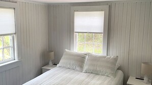 A bedroom with a large bed, two bedside tables, and two windows with blinds. - Cozy Cape Cottage with Beach Access (Eastham)