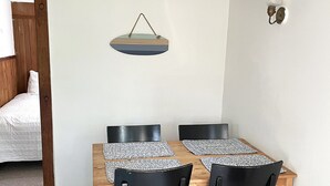 A dining area with a wooden table and four black chairs, a hanging light fixture, and a bed in the background. - Cozy Cape Cottage with Beach Access (Eastham)