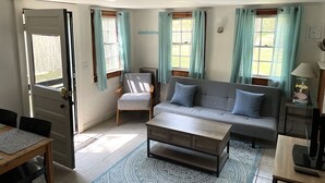 A living room with a sofa, armchair, coffee table, and a dining table. - Cozy Cape Cottage with Beach Access (Eastham)