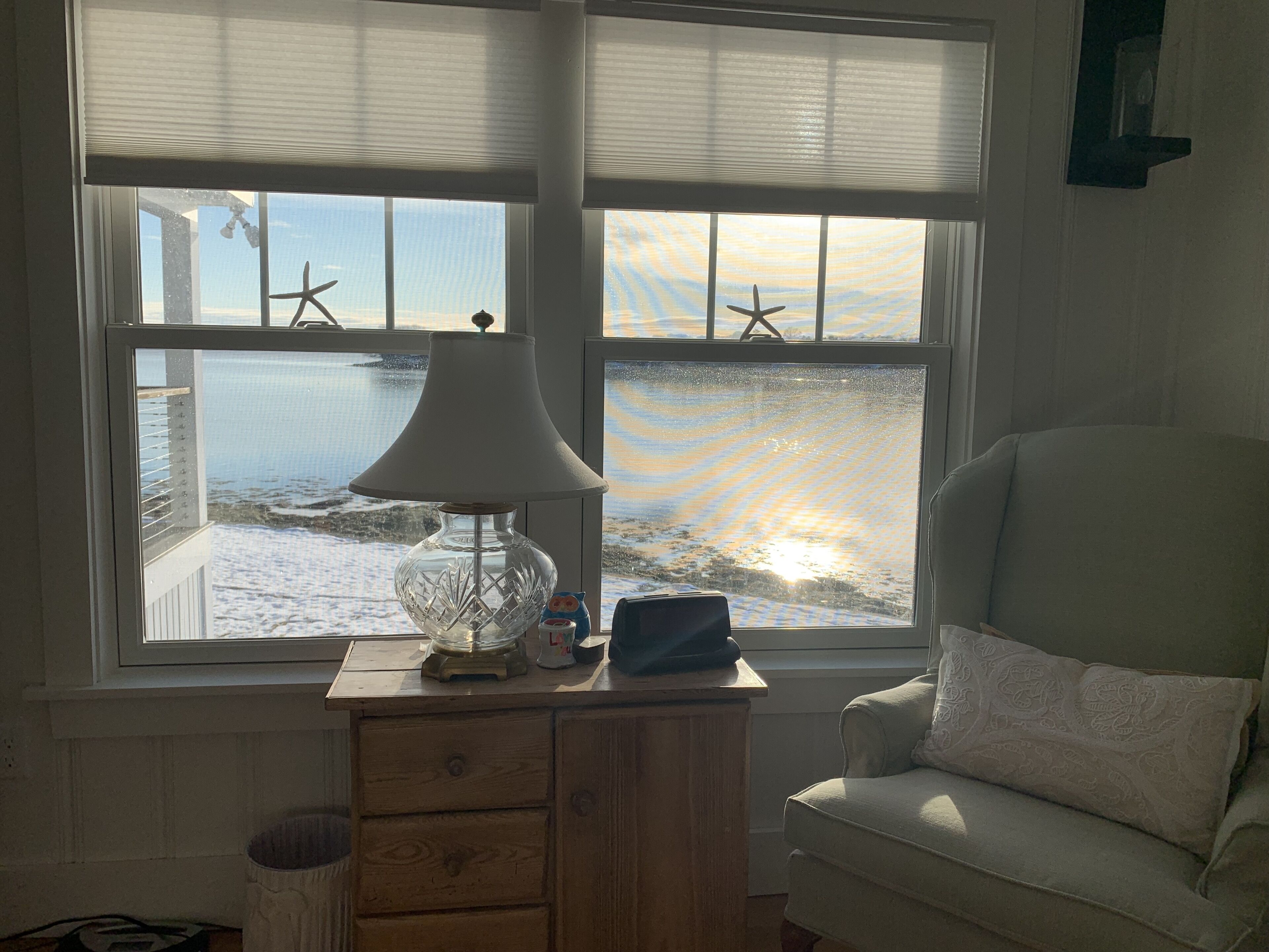 A room with a view of the sea through large windows, a lamp on a wooden table, and a chair with a patterned cushion.