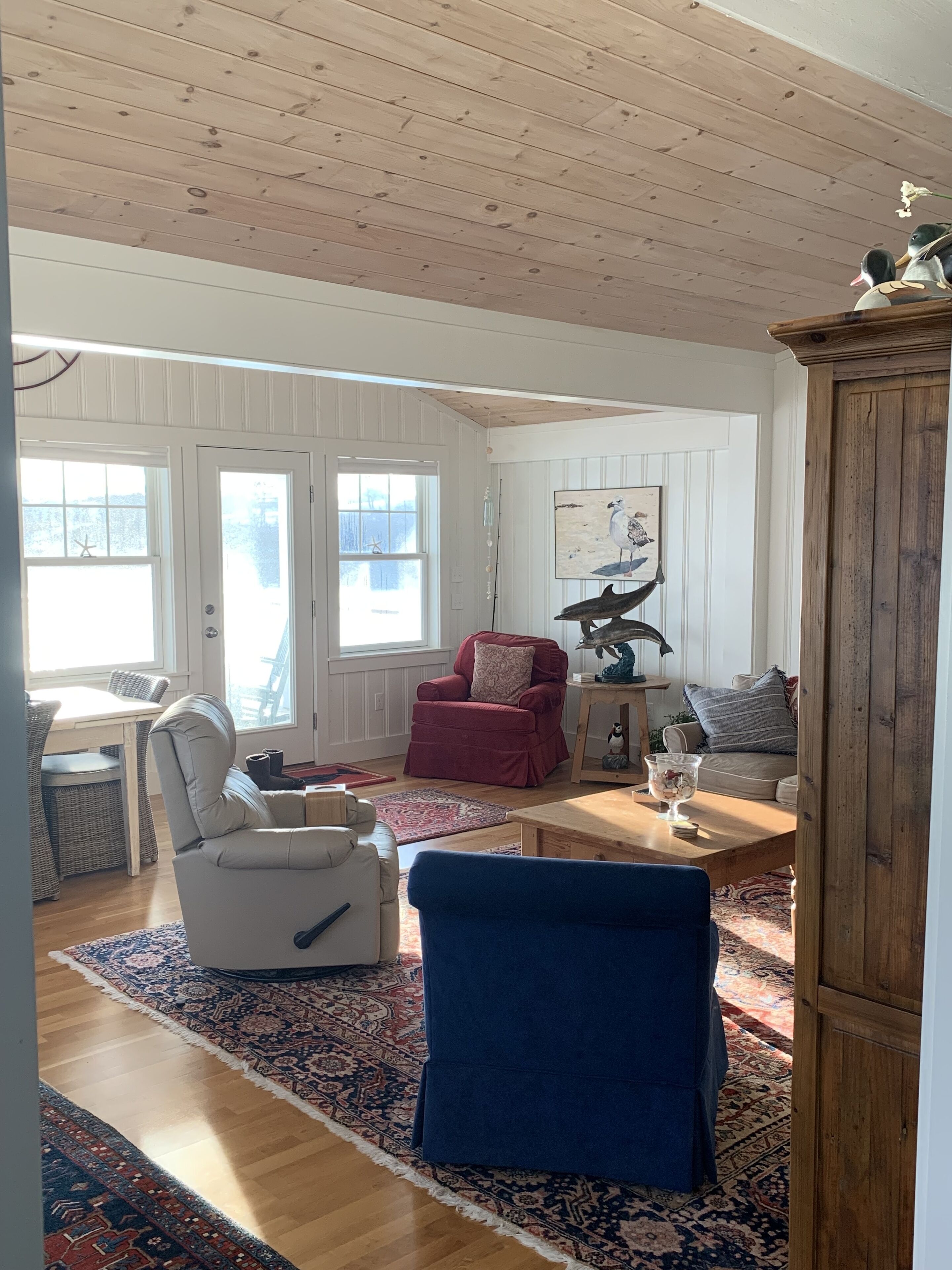 A cozy living room with a wooden ceiling, a red armchair, a white armchair, a blue armchair, a wooden coffee table, and a rug.