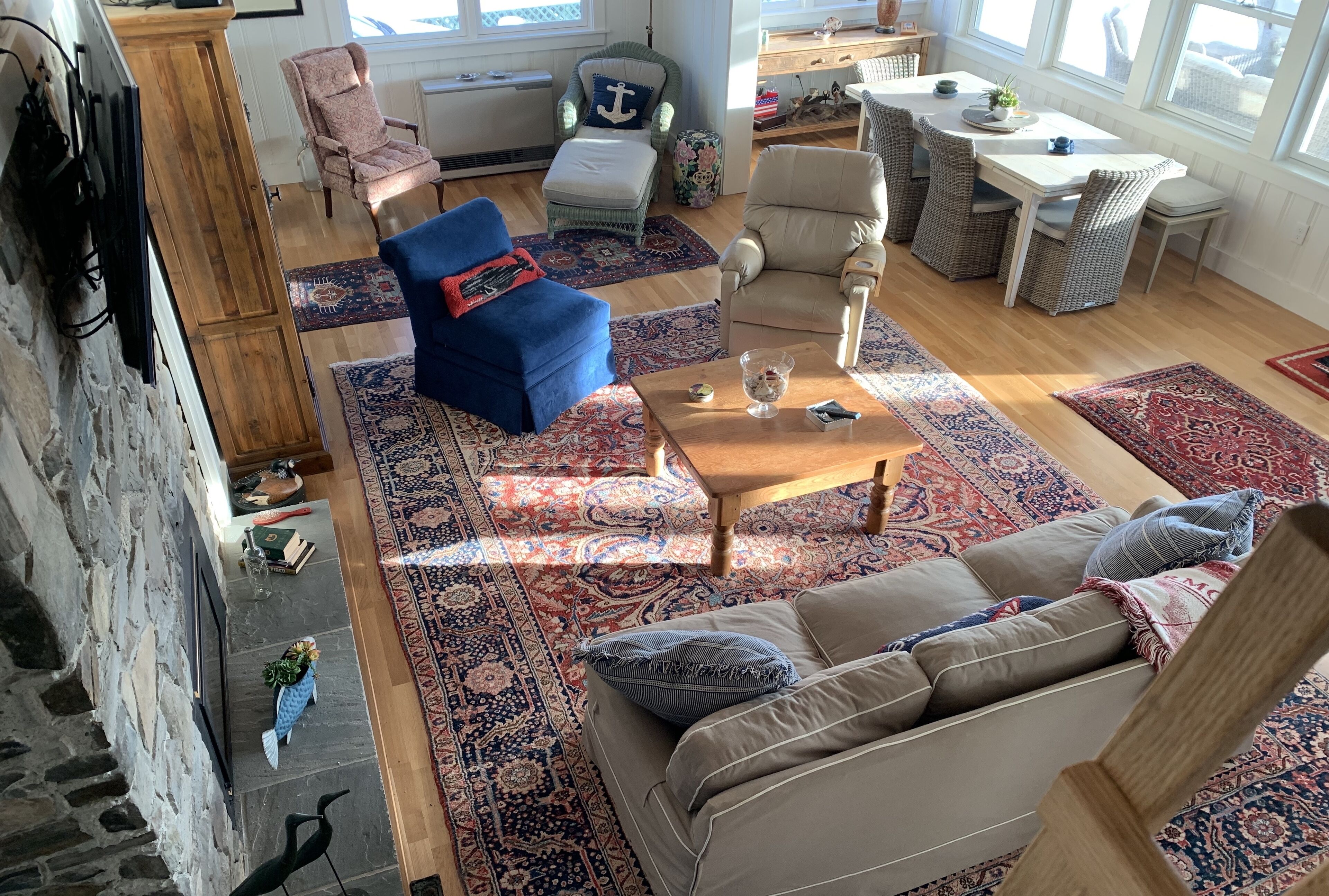 Un salon spacieux avec un fauteuil bleu, un canapé beige et une table basse en bois. On y trouve plusieurs chaises et une table à manger avec des chaises. La pièce est dotée de grandes fenêtres et d’un mur en pierre.