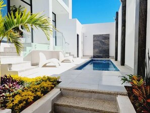 Et moderne poolområde med en klar blå pool, hvide trin ned til poolen og potteplanter langs kanten.