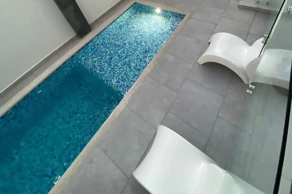 An indoor swimming pool with a tiled floor, a small garden area with plants, and white lounge chairs.