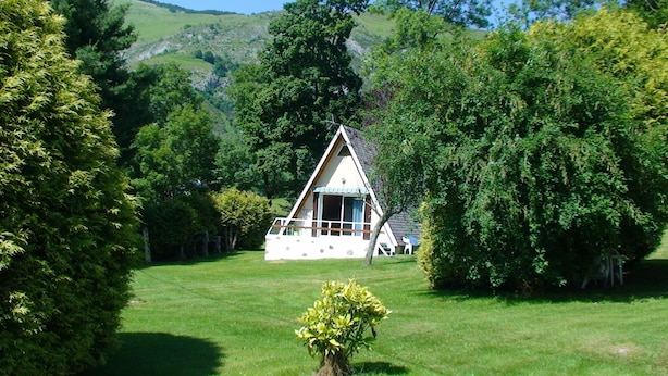 A white, triangular-roofed cabin surrounded by lush greenery and trees, with a clear view of the surrounding landscape.