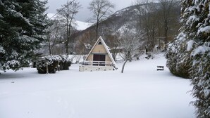A snow-covered landscape with a triangular-roofed building, surrounded by snow-laden trees and mountains in the background.