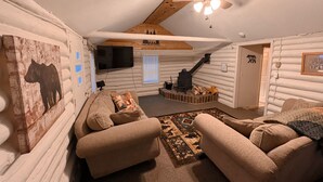 A cozy log cabin interior with a sofa, a ceiling fan, a fireplace, and a mounted television.