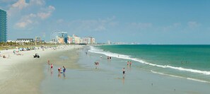 A beach with people swimming and walking along the shore, a Ferris wheel, and a row of buildings in the background. - 8/5/23-8/11/23 Beach Vacation (North Myrtle Beach)